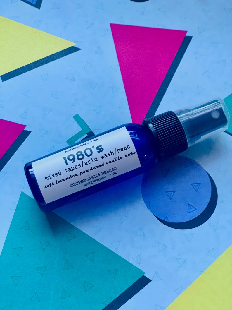 May include: A blue glass bottle with a black spray nozzle and white text that reads "1980's mixed tapes/acid wash/neon soft lavender/powdered vanilla/rose" and "DISTILLED WATER, ESSENTIAL & FRAGRANCE OILS, NATURAL PRESERVATIVE 1 OZ".