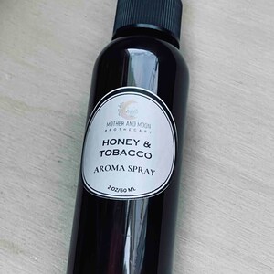 May include: A black glass bottle with a white label that reads "Mother and Moon Apothecary Honey & Tobacco Aroma Spray 2 oz/60 ml".
