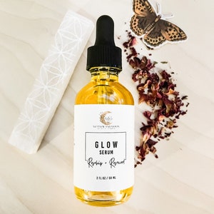 Peut inclure: Une bouteille en verre transparent avec un compte-gouttes noir rempli d'un liquide jaune. L'&eacute;tiquette indique "GLOW SERUM Rosehip + Rosewater 2 FL OZ / 59 ML" et pr&eacute;sente un croissant de lune et une &eacute;toile. La bouteille est entour&eacute;e de p&eacute;tales de rose s&eacute;ch&eacute;s et d'un autocollant de papillon.