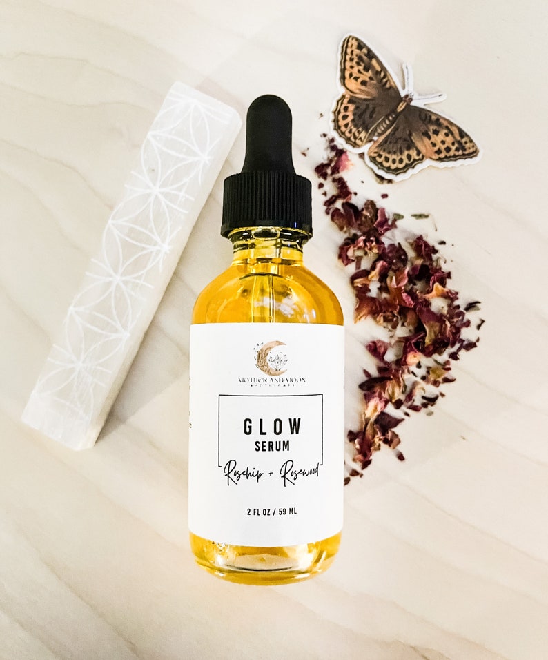 Peut inclure: Une bouteille en verre transparent avec un compte-gouttes noir rempli d'un liquide jaune. L'&eacute;tiquette indique "GLOW SERUM Rosehip + Rosewater 2 FL OZ / 59 ML" et pr&eacute;sente un croissant de lune et une &eacute;toile. La bouteille est entour&eacute;e de p&eacute;tales de rose s&eacute;ch&eacute;s et d'un autocollant de papillon.
