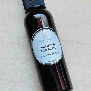 May include: A dark brown glass bottle with a black spray nozzle and a white label that reads "Mother and Moon Apothecary Honey & Tobacco Aroma Spray 2 oz/60 ml."