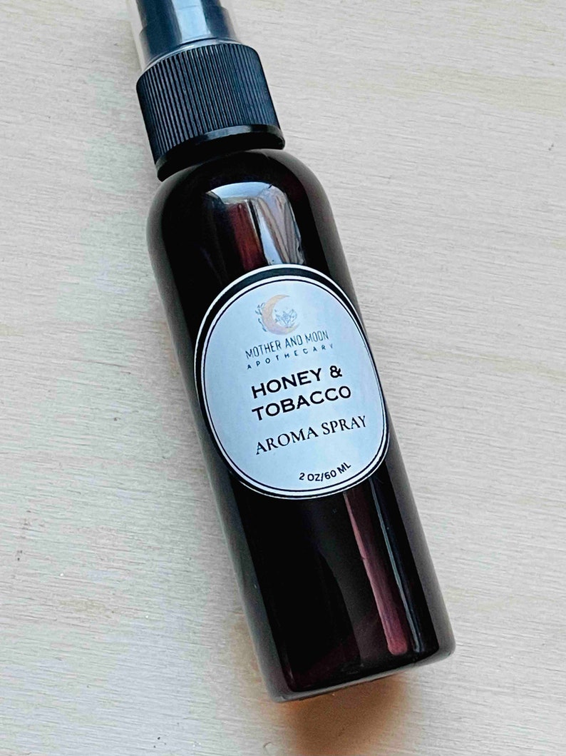 May include: A dark brown glass bottle with a black spray nozzle and a white label that reads "Mother and Moon Apothecary Honey & Tobacco Aroma Spray 2 oz/60 ml."