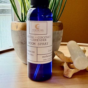 May include: A blue glass room spray bottle with a black cap, labeled "SANTAL - COCONUT - LAVENDER ROOM SPRAY." The bottle is 2 oz. and sits on a wooden tray with a white crystal and decorative wooden pieces. Two potted plants are in the background.