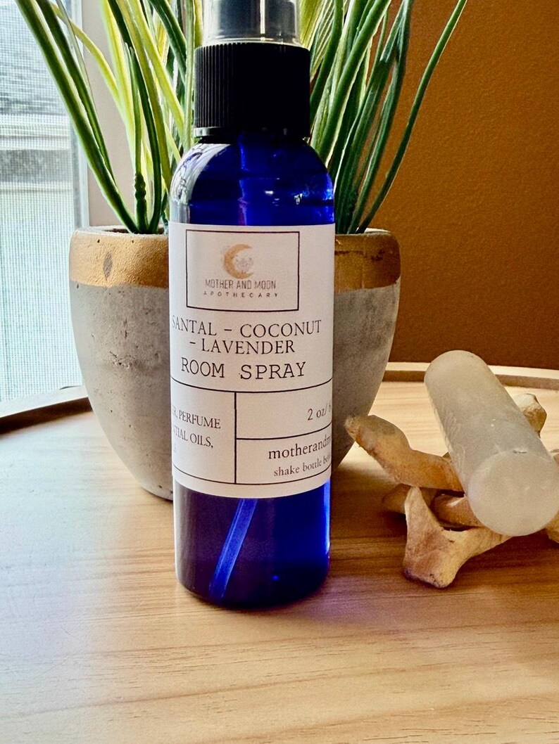 May include: A blue glass room spray bottle with a black cap, labeled "SANTAL - COCONUT - LAVENDER ROOM SPRAY." The bottle is 2 oz. and sits on a wooden tray with a white crystal and decorative wooden pieces. Two potted plants are in the background.