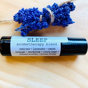 May include: A black and silver roller bottle with a white label that reads "SLEEP aromatherapy blend vetiver + lavender + sweet orange + marjoram + clary sage + frankincense + ylang ylang".