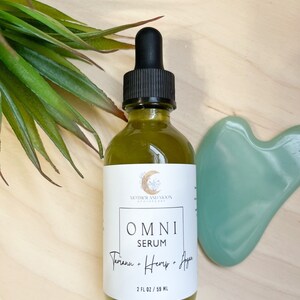 May include: A clear glass bottle with a black dropper top filled with a green liquid. The bottle has a white label with the text "MOTHER AND MOON BOTANICALS" and "OMNI SERUM Tamanu + Hemp + Agave 2 FL OZ / 59 ML".