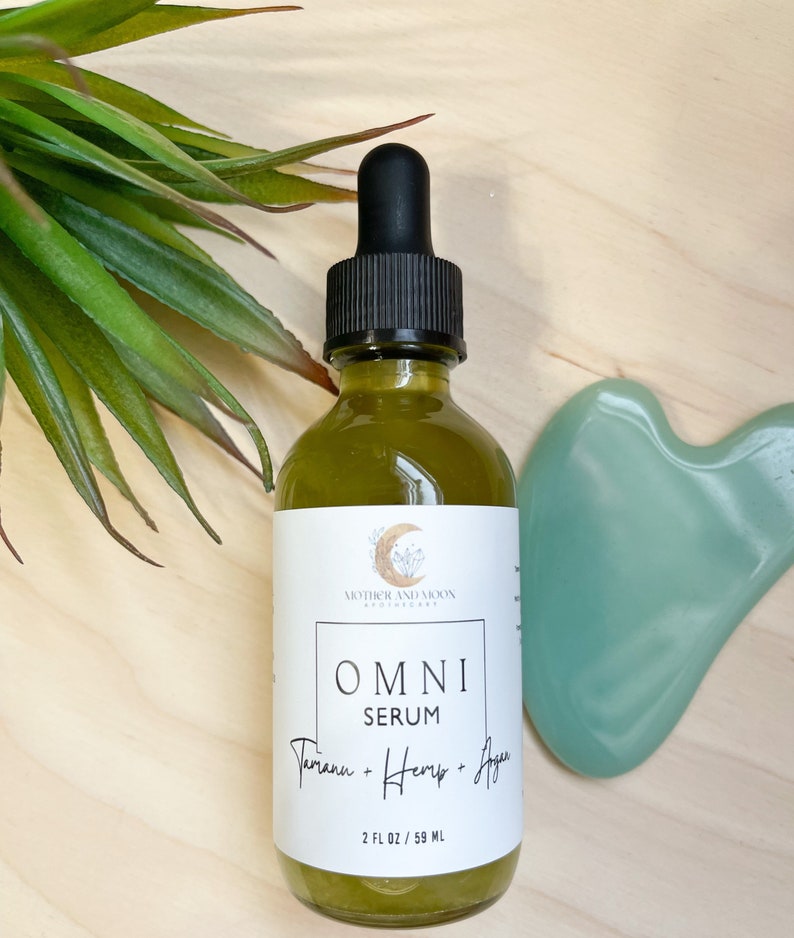 May include: A clear glass bottle with a black dropper top filled with a green liquid. The bottle has a white label with the text "MOTHER AND MOON BOTANICALS" and "OMNI SERUM Tamanu + Hemp + Agave 2 FL OZ / 59 ML".