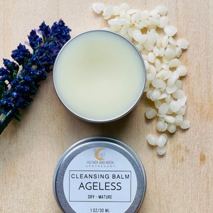May include: A silver tin with a white label that reads "MOTHER AND MOON APOTHECARY CLEANSING BALM AGELESS DRY - MATURE 1 OZ/30 ML". The tin is sitting on a wooden surface with white wax beads scattered around it.