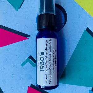 May include: A blue glass bottle with a black spray top. The label reads "1980's mixed tapes/acid wash/neon soft lavender/powdered vanilla/rosc" and "motherandmoon.com". The bottle is sitting on a colorful background.