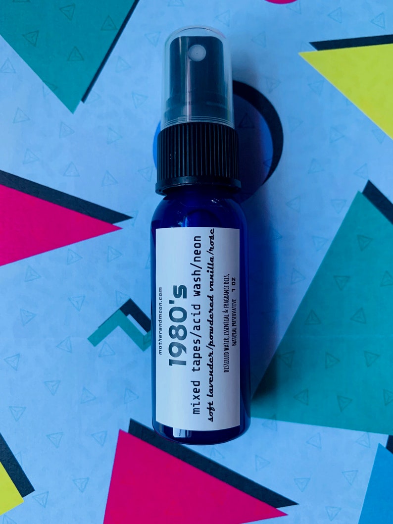 May include: A blue glass bottle with a black spray top. The label reads "1980's mixed tapes/acid wash/neon soft lavender/powdered vanilla/rosc" and "motherandmoon.com". The bottle is sitting on a colorful background.