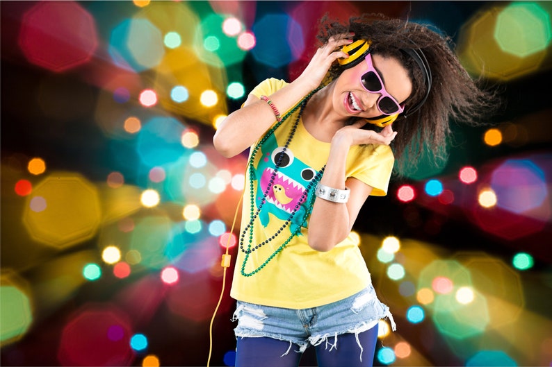 May include: A young woman with long brown hair is wearing yellow and blue clothing, pink sunglasses, and yellow headphones. She is smiling and listening to music. The background is a blur of colorful lights.