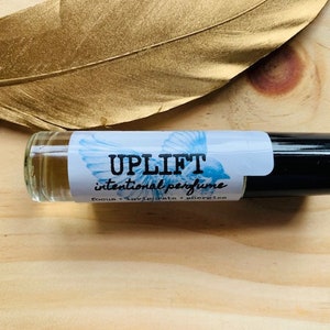 May include: A clear glass roll-on bottle with a black cap. The label is white with blue accents and says "UPLIFT intentional perfume  romans + lavendar + gingerly"