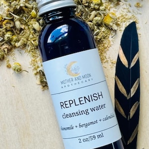 May include: A blue glass bottle with a silver lid, labeled "REPLENISH cleansing water chamomile + bergamot + calendula 2 oz/59 ml" and "MOTHER AND MOON APOTHECARY".
