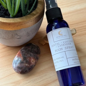 May include: A blue room spray bottle with a black spray top, labeled "SANTAL - COCONUT - LAVENDER ROOM SPRAY." A decorative rock and a potted plant are next to the bottle. The bottle contains 2 oz of product.
