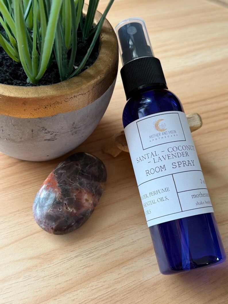May include: A blue room spray bottle with a black spray top, labeled "SANTAL - COCONUT - LAVENDER ROOM SPRAY." A decorative rock and a potted plant are next to the bottle. The bottle contains 2 oz of product.