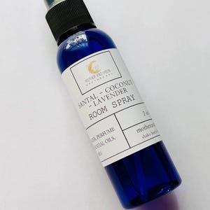 May include: A blue glass room spray bottle with a black spray nozzle. The label reads "SANTAL - COCONUT - LAVENDER ROOM SPRAY" and "MOTHER AND MOON APOTHECARY". The bottle contains 2 oz of product.
