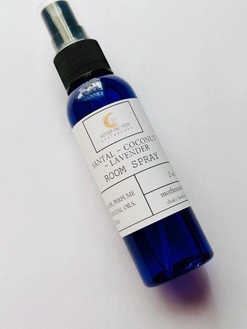 May include: A blue glass room spray bottle with a black spray nozzle. The label reads "SANTAL - COCONUT - LAVENDER ROOM SPRAY" and "MOTHER AND MOON APOTHECARY". The bottle contains 2 oz of product.