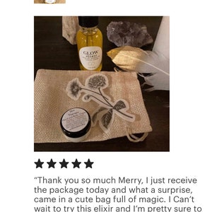 Peut inclure: Un petit sac en toile de jute avec un motif floral, contenant un petit pot de cr&egrave;me de soin pour la peau et un flacon en verre d'&eacute;lixir de soin du visage. Le flacon porte une &eacute;tiquette noire avec les mots "Glow Elixir" imprim&eacute;s dessus. L'image comprend &eacute;galement une plume et un cristal.