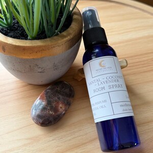 May include: A blue room spray bottle with a black spray nozzle, labeled "SANTAL - COCONUT - LAVENDER ROOM SPRAY." A small potted plant and a polished stone are next to the bottle on a wooden surface.