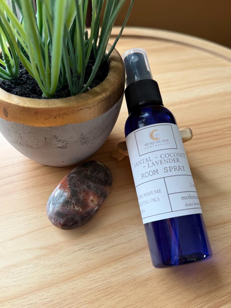 May include: A blue room spray bottle with a black spray nozzle, labeled "SANTAL - COCONUT - LAVENDER ROOM SPRAY." A small potted plant and a polished stone are next to the bottle on a wooden surface.