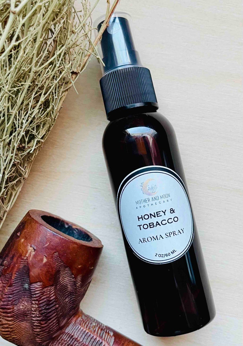 May include: A brown glass bottle with a black spray top and a white label that reads "Mother and Moon Apothecary Honey & Tobacco Aroma Spray 2 oz/60 ml".