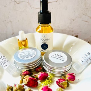 May include: A white plate with five small bottles and jars containing various beauty products. The products include a face cream, hand and body cream, aroma spray, and a serum. The labels on the products include the names "Nourish Elixir", "Luxe Face Cream", "Skin Therapy Hand & Body Cream", "No. 31 Aroma Spray", and "Rytelle Aroma Spray".