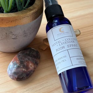 May include: A blue room spray bottle with a black cap and a white label that reads "SANTAL - COCONUT - LAVENDER ROOM SPRAY." A potted plant and a polished stone are next to the bottle. The bottle is 2 fluid ounces.