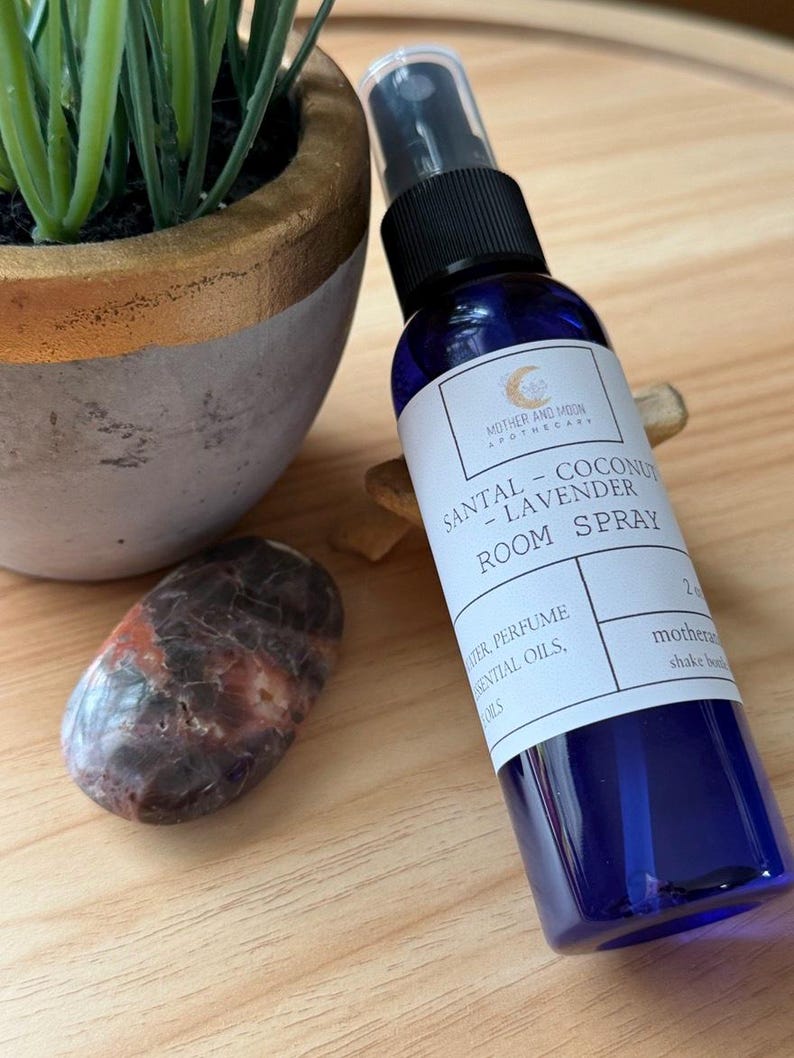 May include: A blue room spray bottle with a black cap and a white label that reads "SANTAL - COCONUT - LAVENDER ROOM SPRAY." A potted plant and a polished stone are next to the bottle. The bottle is 2 fluid ounces.