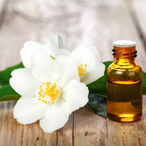 May include: A small amber glass bottle of jasmine essential oil with a white cap, sitting on a wooden surface with white jasmine flowers and green leaves.