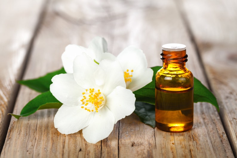 May include: A small amber glass bottle of jasmine essential oil with a white cap, sitting on a wooden surface with white jasmine flowers and green leaves.