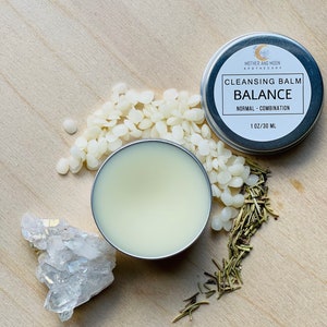 May include: A silver tin of cleansing balm labeled "Cleansing Balm Balance" by Mother and Moon Apothecary. The tin is 1 oz / 30 ml and is for normal to combination skin. The balm is in a small silver container with a white lid. There are white wax beads and dried rosemary sprigs scattered around the tin.