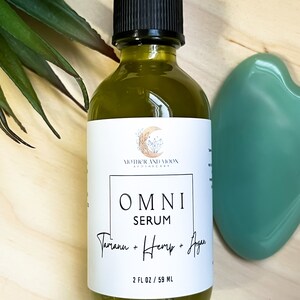 May include: A glass bottle of green serum labeled "OMNI SERUM Tamanu + Hemp + Agan" with a white label and black text. The bottle is 2 fl oz / 59 ml and is sitting on a wooden surface.