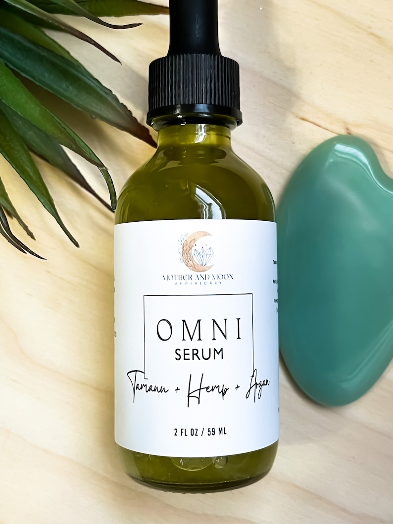 May include: A glass bottle of green serum labeled "OMNI SERUM Tamanu + Hemp + Agan" with a white label and black text. The bottle is 2 fl oz / 59 ml and is sitting on a wooden surface.