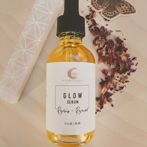 Peut inclure: Une bouteille en verre de s&eacute;rum &eacute;tiquet&eacute;e "Glow Serum Rosehip + Rosewater" avec un bouchon compte-gouttes noir. La bouteille est pos&eacute;e sur une surface en bois avec des p&eacute;tales de rose s&eacute;ch&eacute;s et un cristal blanc.