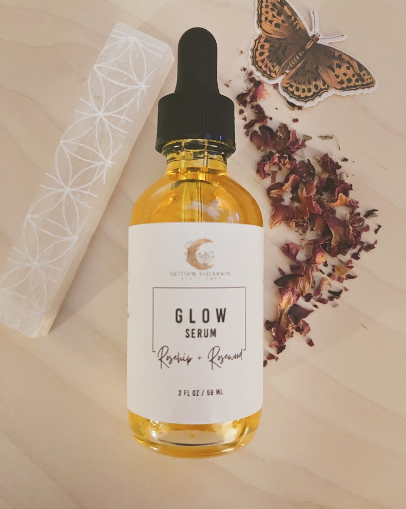 Peut inclure: Une bouteille en verre de s&eacute;rum &eacute;tiquet&eacute;e "Glow Serum Rosehip + Rosewater" avec un bouchon compte-gouttes noir. La bouteille est pos&eacute;e sur une surface en bois avec des p&eacute;tales de rose s&eacute;ch&eacute;s et un cristal blanc.