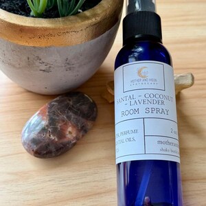 May include: A blue room spray bottle with a black spray top, labeled "SANTAL - COCONUT - LAVENDER ROOM SPRAY." The bottle is next to a polished stone and a small potted plant. The bottle contains 2 fl oz.
