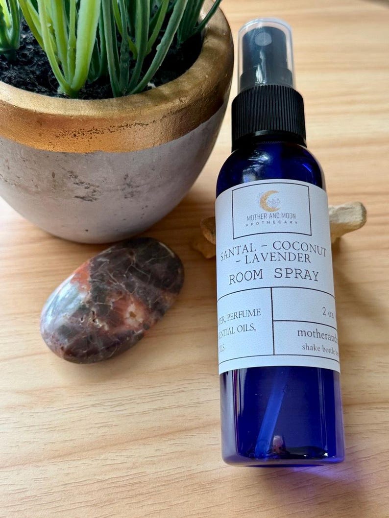 May include: A blue room spray bottle with a black spray top, labeled "SANTAL - COCONUT - LAVENDER ROOM SPRAY." The bottle is next to a polished stone and a small potted plant. The bottle contains 2 fl oz.
