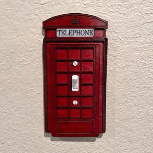 Puede incluir: Una cubierta de interruptor de luz de madera roja con forma de cabina telefónica británica. La palabra "TELEPHONE" está en la parte superior. La cubierta tiene un interruptor de luz blanco. Está montada en una pared texturizada blanquecina.