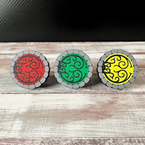 May include: Three round decorative wooden buttons with a gray border and a black edge. Each button features a different color: red, green, and yellow, with a black swirling design in the center. The buttons are displayed on a wooden surface.
