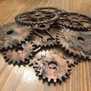 Puede incluir: Un conjunto de ocho engranajes metálicos de diferentes tamaños, pintados en un acabado negro y cobre desgastado. Los engranajes están dispuestos en un patrón aleatorio sobre una superficie de madera.