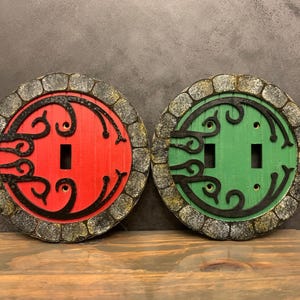 May include: Two decorative light switch covers with a stone-like design. One cover is red with black details and the other is green with black details. Both covers feature a stylized design of a door with a swirling pattern.