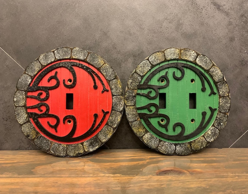 May include: Two decorative light switch covers with a stone-like design. One cover is red with black details and the other is green with black details. Both covers feature a stylized design of a door with a swirling pattern.