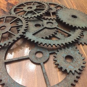 May include: A set of six rustic metal gears with a distressed, aged finish. The gears are arranged in a circular pattern, with the largest gear in the center. The gears are painted in shades of brown and green, giving them a vintage look.