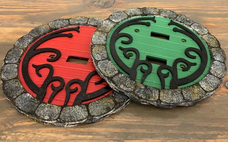 May include: Two decorative light switch covers with a stone-like border. One cover is red with black detailing and the other is green with black detailing.