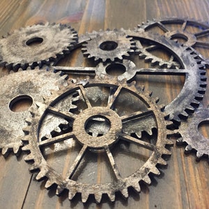 Puede incluir: Una composición decorativa de engranajes de madera cortados con precisión, dispuestos en capas para crear profundidad. Los engranajes presentan un acabado plateado y marrón envejecido, creando una estética rústica e industrial. Sobre fondo de madera oscura.