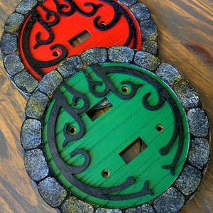 May include: Two round light switch covers with a stone-like border. One cover is red with black detailing and the other is green with black detailing.