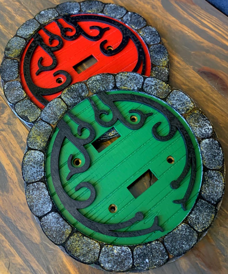 May include: Two round light switch covers with a stone-like border. One cover is red with black detailing and the other is green with black detailing.