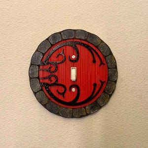 May include: A round light switch cover featuring a red door design with a black stone-like border. The door has a black swirl design and a white light switch.