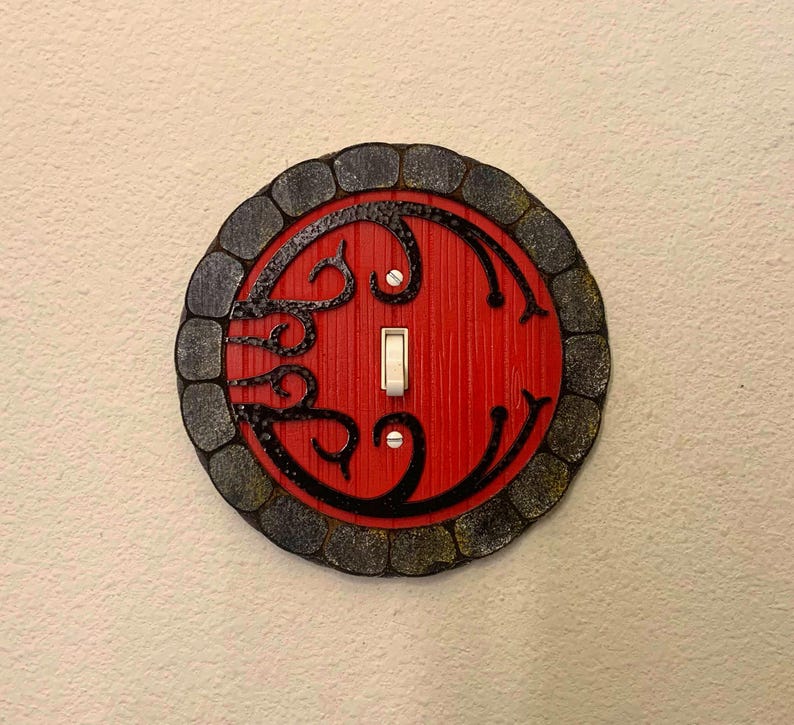 May include: A round light switch cover with a red door design and black stone-like border. The door has a black swirl design and a white light switch.