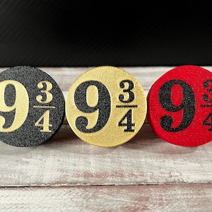 May include: Three round decorative objects with the number "9 3/4" in a bold font. One is black with gold numbers, one is gold with black numbers, and one is red with black numbers. They are arranged on a light wood surface.